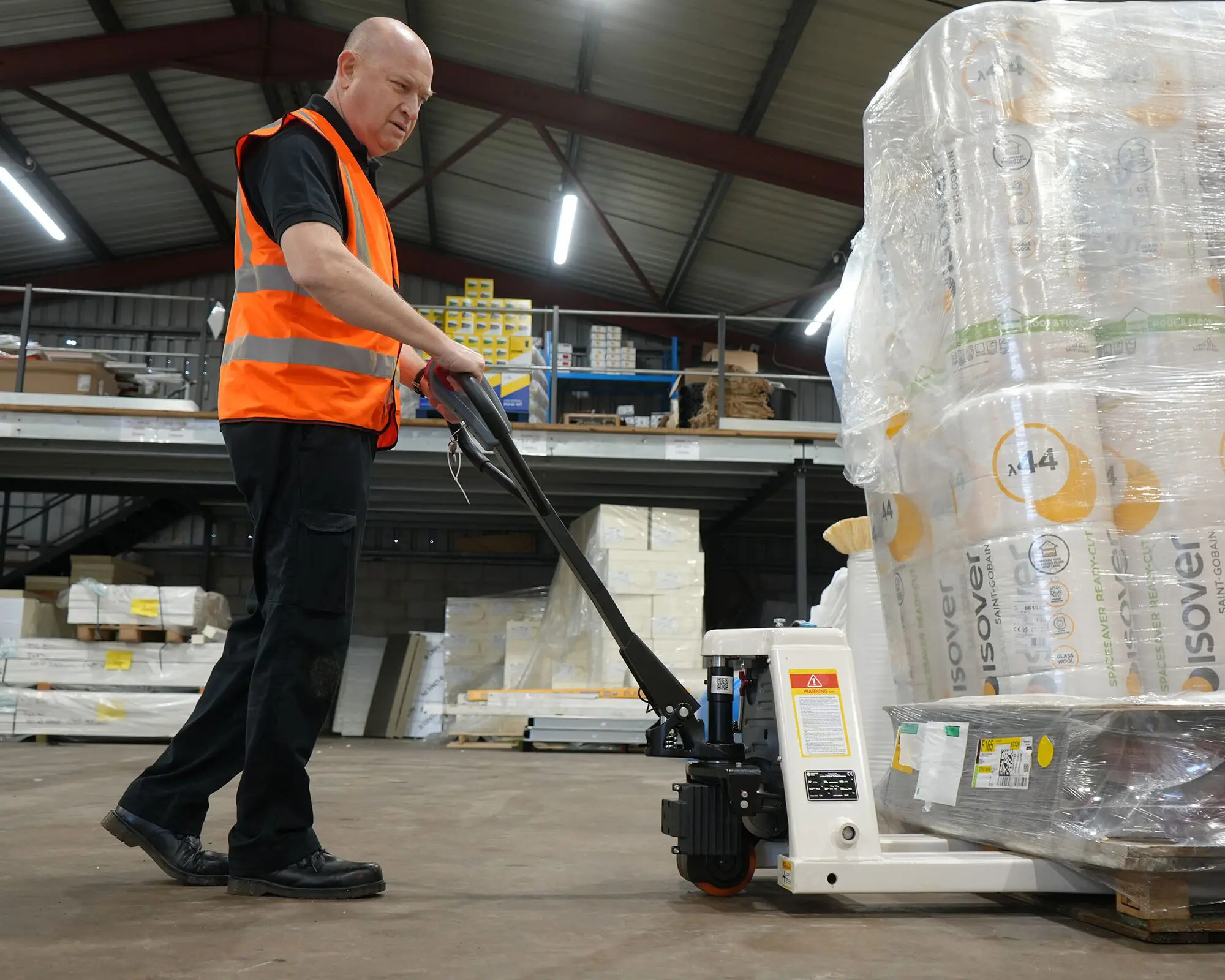 A warehouse worker in a safety vest using a pallet truck to move boxes of materials.