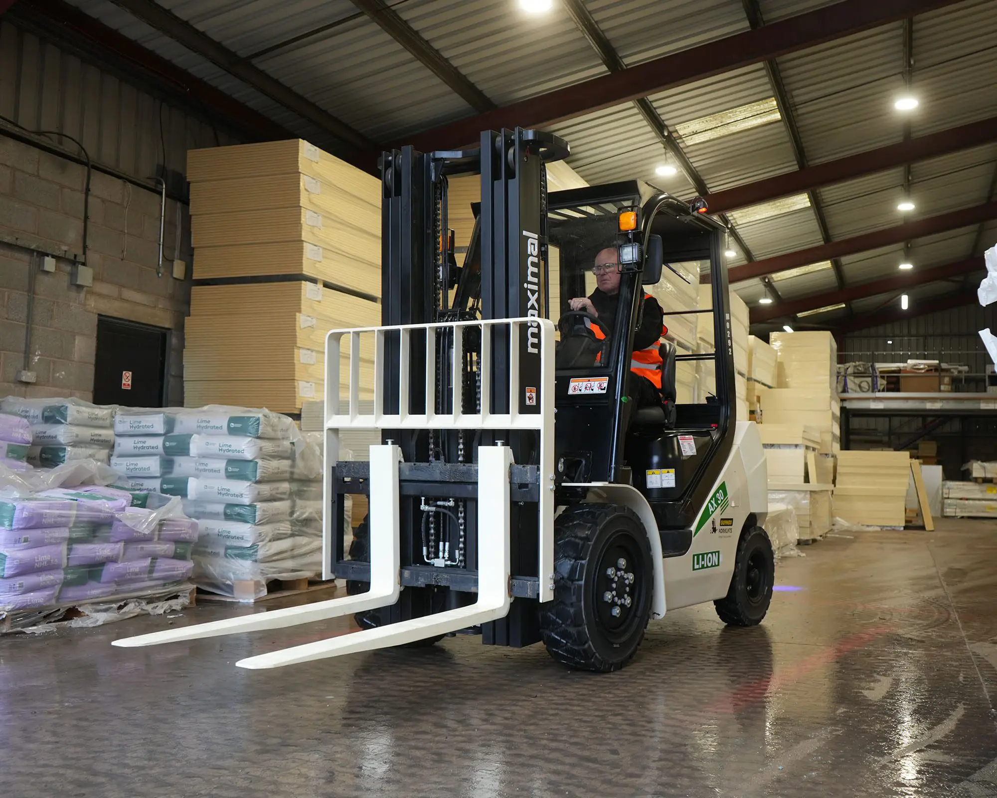 A forklift operator maneuvering a forklift with long forks inside a warehouse filled with stacked materials.