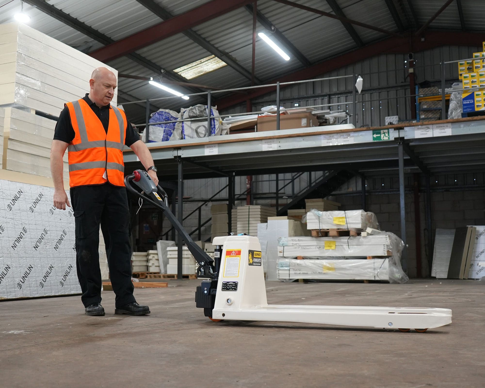 A warehouse worker in a safety vest using a pallet truck in a storage facility with shelves and goods in the background.