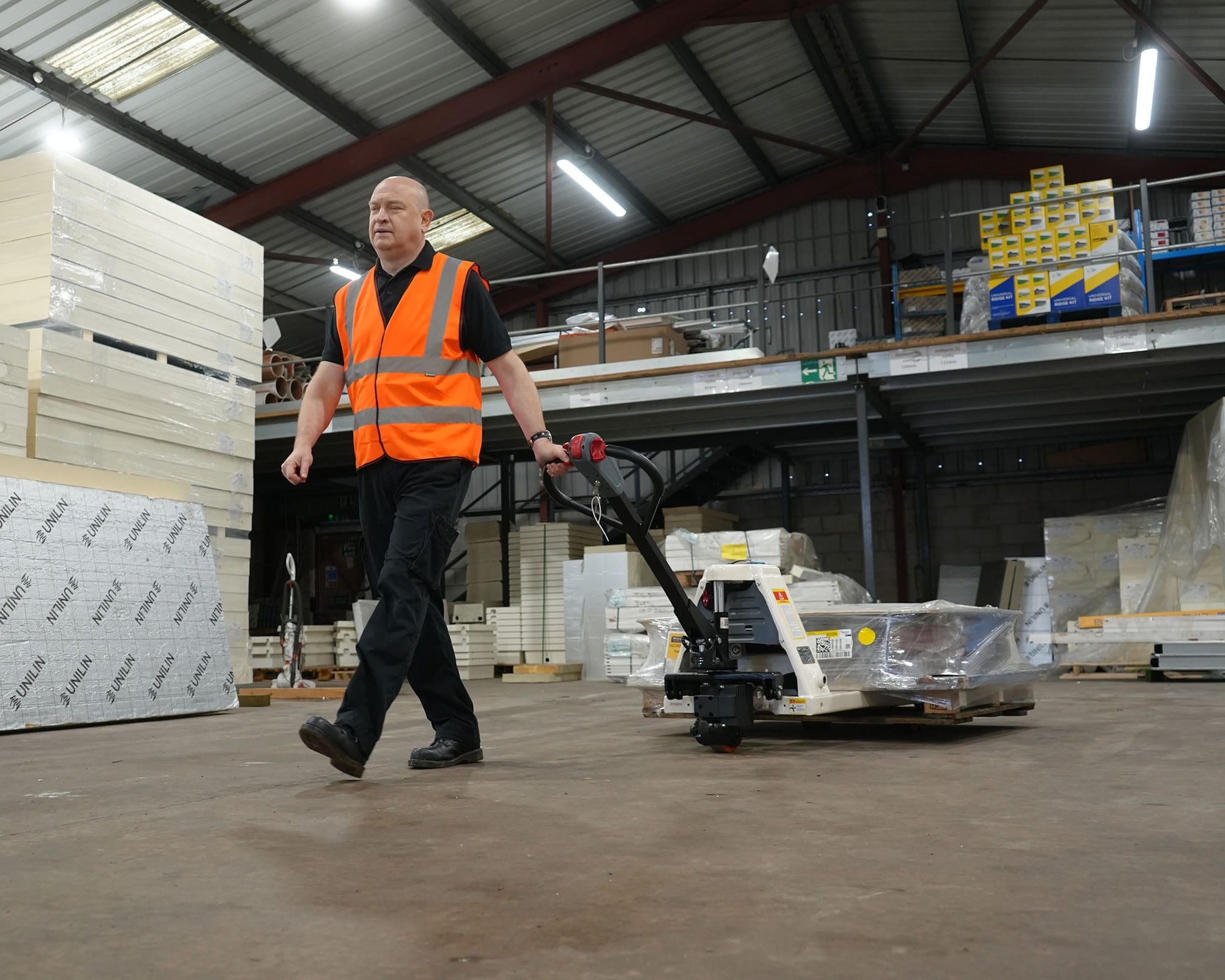 A warehouse worker in a safety vest operating a pallet truck in a storage area filled with goods.