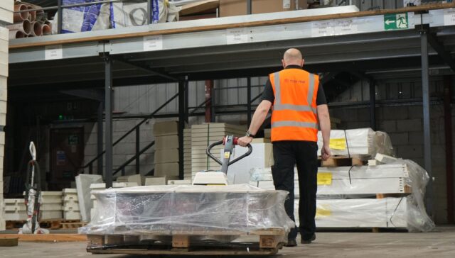 A warehouse employee in a safety vest moving a pallet loaded with materials using a pallet truck.