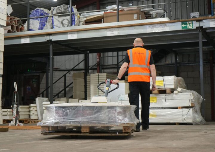 A warehouse employee in a safety vest moving a pallet loaded with materials using a pallet truck.