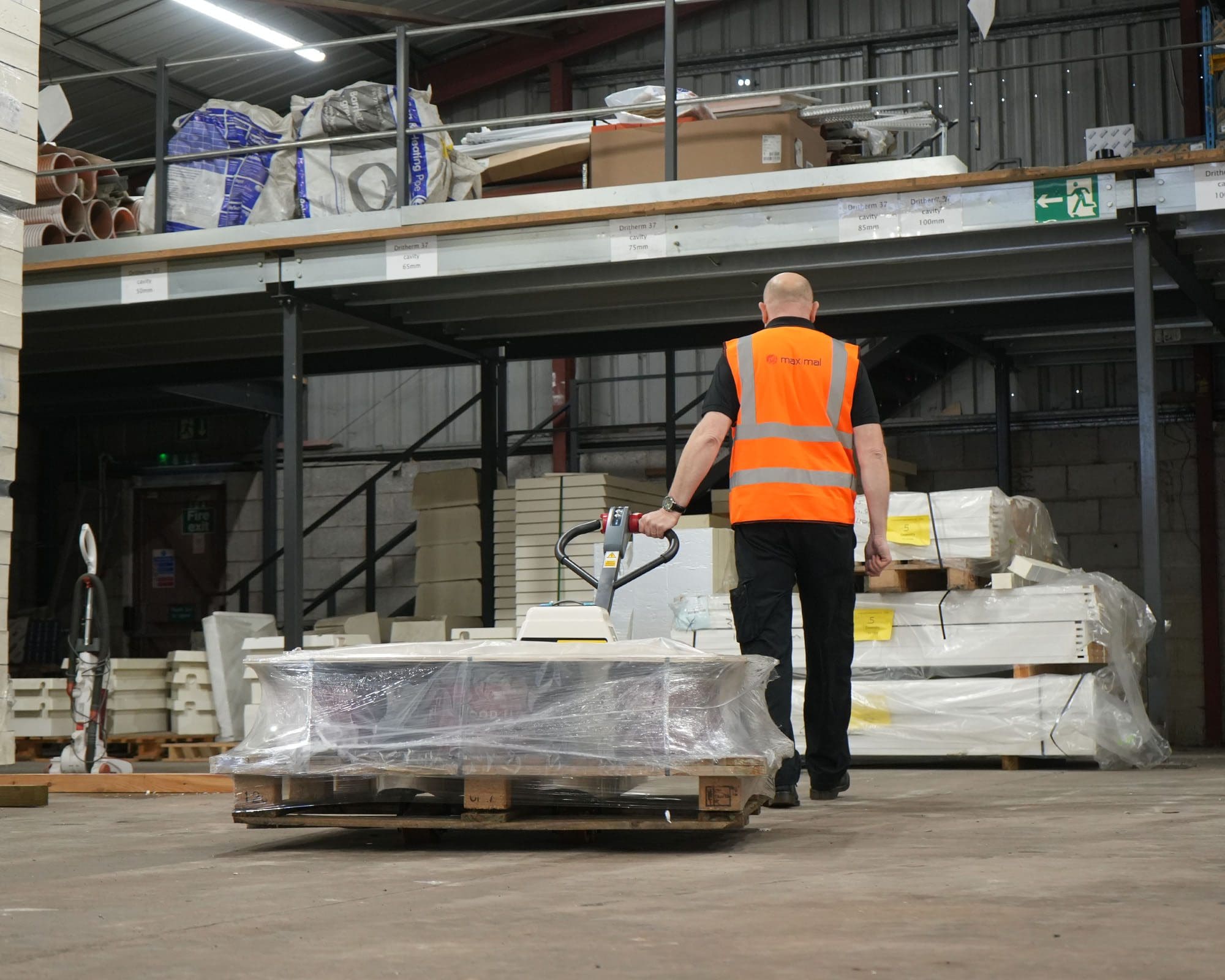 Warehouse Worker Transporting Goods with Pallet Truck A warehouse employee in a safety vest moving a pallet loaded with materials using a pallet truck.